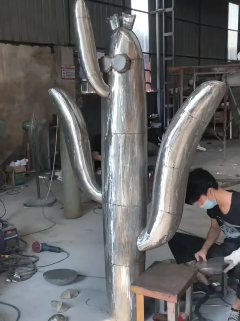 A skilled artisan fabricating a large, polished stainless steel metal cactus sculpture in a workshop. This sleek cactus sculpture metal piece offers surreal clarity and absolute permanence, perfectly replacing fragile organic planting to redefine low-maintenance luxury in modern arid courtyards.