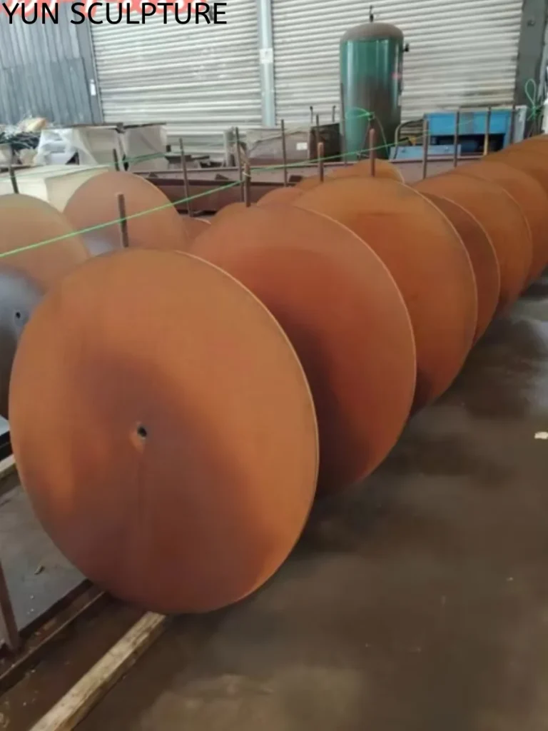 A neat row of large, circular Corten steel top plates undergoing factory-controlled pre-oxidation processes in a metal workshop. Through the precise application of chemical catalysts and controlled wet-dry cycles, these components rapidly develop a vibrant, even orange rust layer, stabilizing the surface long before they reach the installation site.