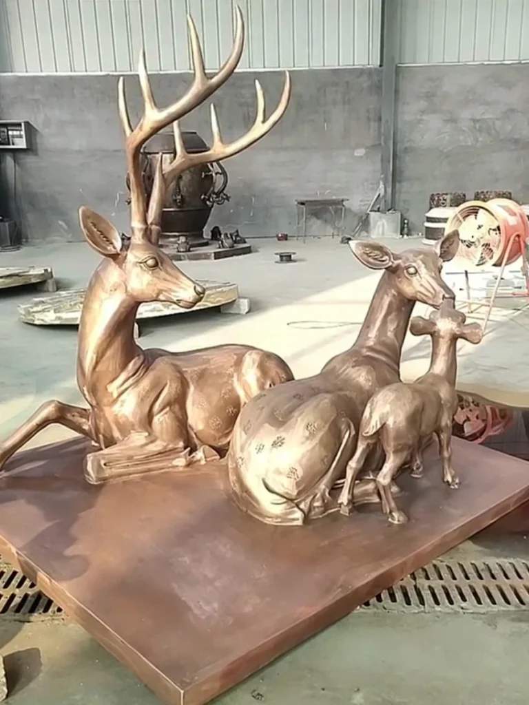 A newly completed bronze deer family sculpture featuring a stag, doe, and fawn resting on a metal base. Freshly treated with chemical patination, the metal exhibits warm golden and rich brown hues, showcasing the elegant aging process that makes bronze the gold standard for landscape art.