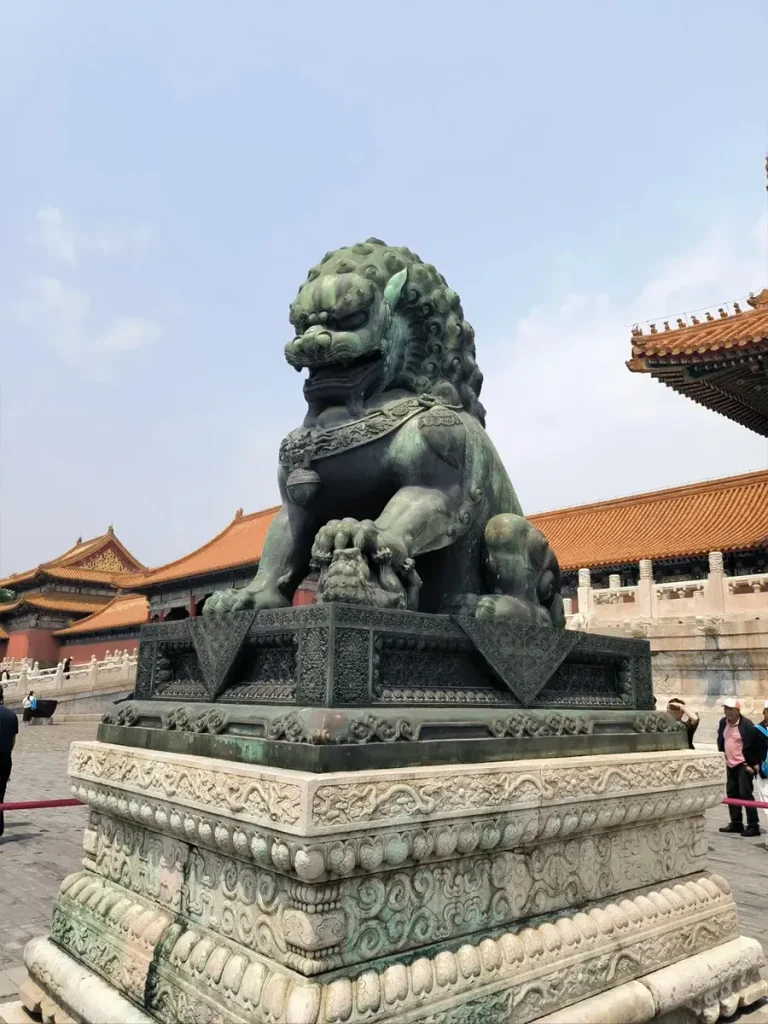 Front-right view of the bronze Foo Dog at the Forbidden City. The sculpture is supported by a magnificent double-layered foundation consisting of an intricate bronze pedestal directly above a massive white marble bottom base, with traditional red buildings in the background.
