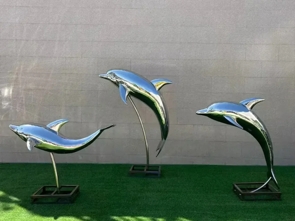 Three forged mirror stainless steel dolphin sculptures with smooth, curved surfaces demonstrating fluid leaping motions against a wall.