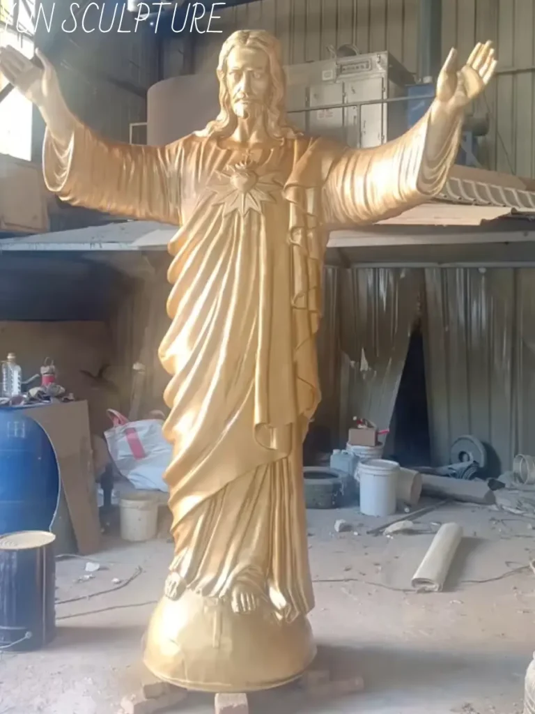 A monumental Bronze Jesus statue gleaming with a raw, golden brass-like finish, standing proudly in a foundry workshop. Newly emerging from the meticulous 9-step Lost-wax casting process, this stunning ecclesiastical commission perfectly demonstrates the incredible capacity for fine detail and the unparalleled strength of true Foundry Bronze designed for centuries of outdoor exposure.