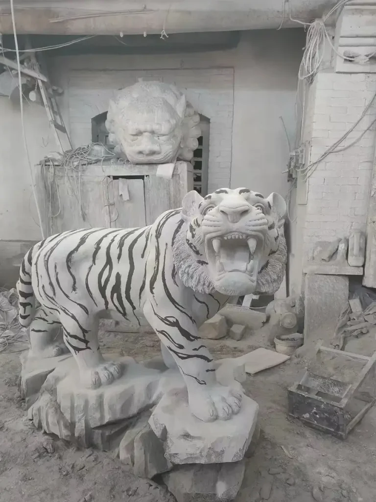 A newly carved life-size bluestone tiger sculpture displayed in a dusty factory workshop. Captured mid-roar on a realistic rock base, the highly detailed stone tiger appears ready to step into a garden landscape, looking as if it has just emerged from the forest edge.