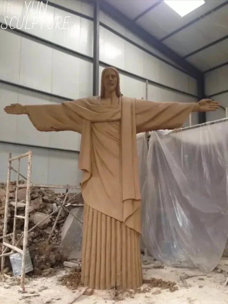 A monumental, full-scale 1:1 clay model of a Risen Christ statue standing in a sculptor's workshop. Showcasing the critical first physical step of custom bronze casting, this towering clay original proves that true ecclesiastical artistry separates custom foundry work from mere mass manufacturing.