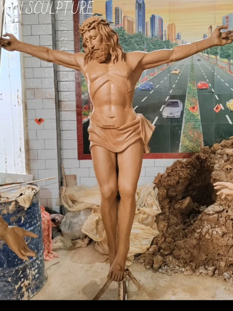 A monumental, life-size clay corpus of a crucified Jesus statue suspended entirely by a temporary metal tripod armature in a bustling workshop. This striking image perfectly illustrates the immense technical challenge of modeling wet clay in a suspended posture—arms extended and weight dropping—without the heavy material collapsing under its own gravity.