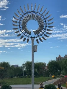 Monumental kinetic stainless steel wind sculpture in dynamic motion, showcasing the mesmerizing Liquid Silver effect. Fabricated from mirror-polished 316L Marine Grade steel elements, the sculpture is set against a backdrop of blue sky, clouds, and an urban landscape, illustrating precision kinetic engineering.