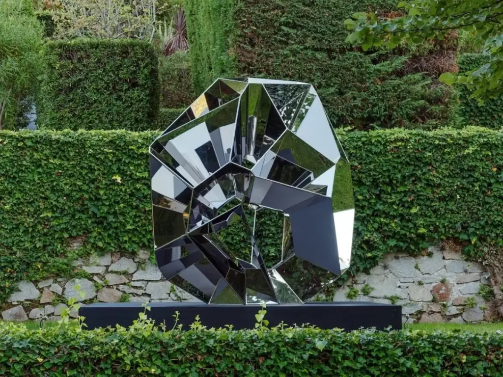 A contemporary geometric metal sculpture placed in a lush garden setting. Constructed from mirror-polished folded panels and intersecting shapes, the artwork perfectly demonstrates how light and shadow act as architectural materials. As natural light hits the multifaceted surface, it creates dynamic layers of depth, sharp highlights, and deep reflections that constantly evolve throughout the day.