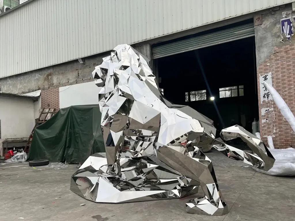 A striking geometric lion sculpture crafted from 316 stainless steel, positioned outside a workshop. The modern faceted design and mirror-polished finish beautifully reflect the surrounding environment, offering a clean, contemporary aesthetic for modern architecture.