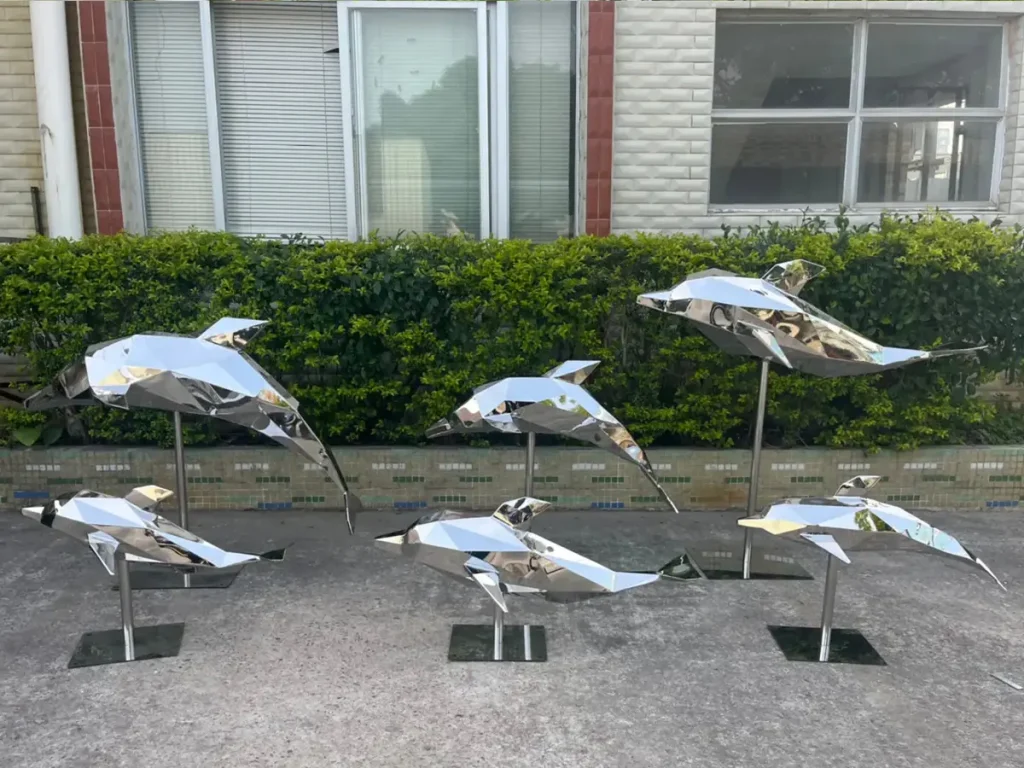 A collection of geometric, faceted mirror stainless steel dolphin sculptures displayed outdoors on metal stands.