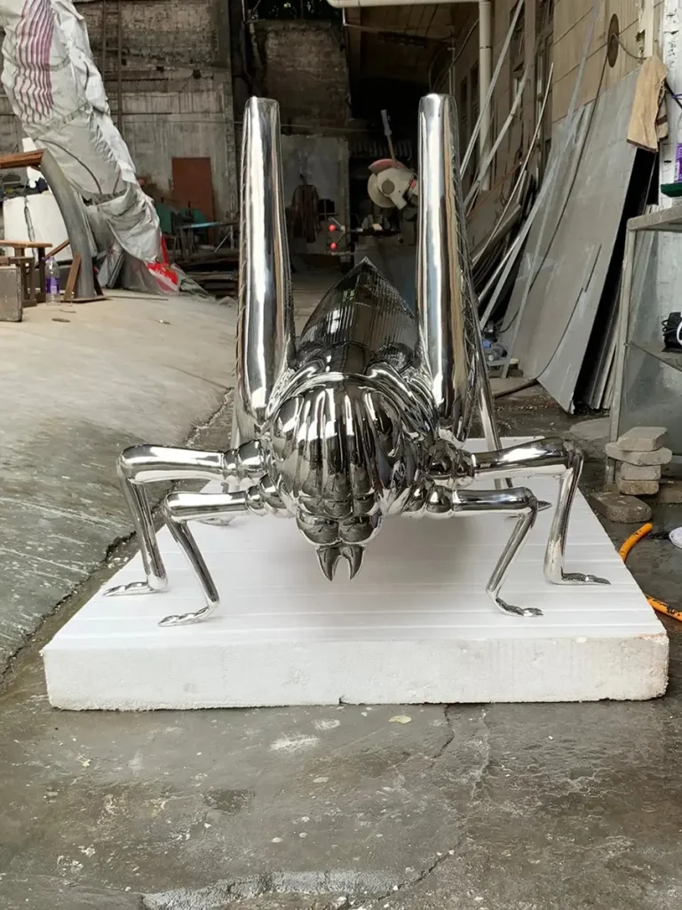 Front view of a giant stainless steel grasshopper sculpture resting on a workshop floor. The flawless mirror-polished finish and oversized, whimsical scale bring fantasy elements to life, proving that modern metal sculpture can completely redefine a garden's atmosphere.
