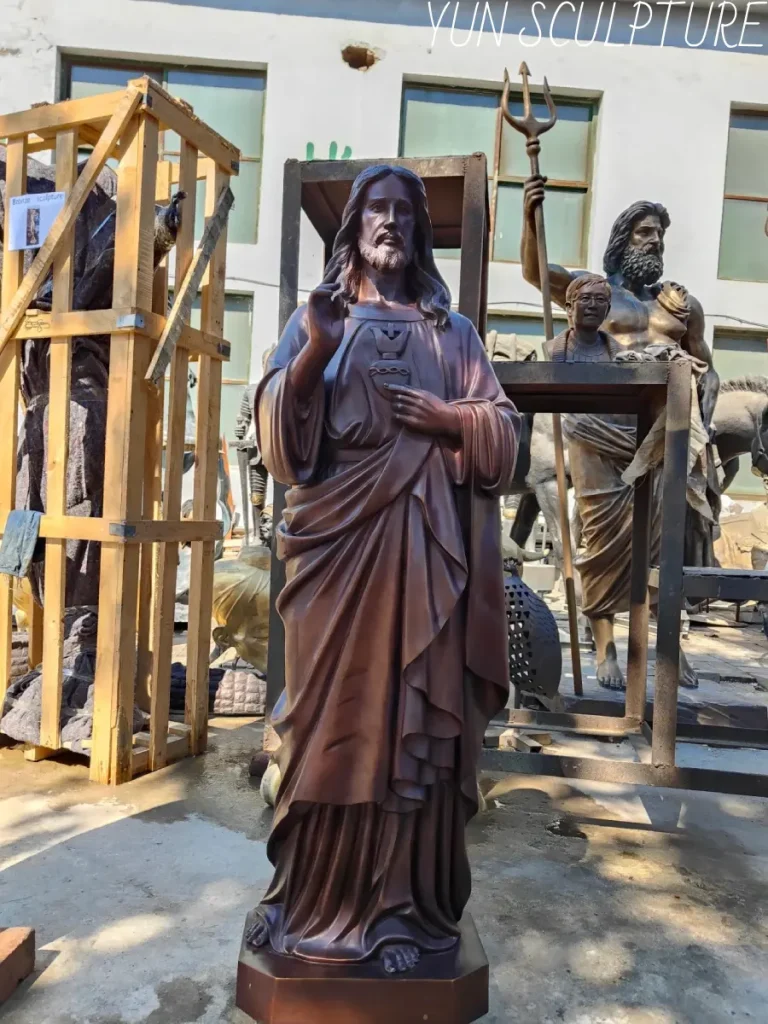 A full-length, majestic bronze divine mercy statue standing in a sculptor's factory yard, ready for global delivery alongside other monumental works in wooden crates. Cast in Grade A Foundry Bronze, this robust ecclesiastical commission is engineered to endure centuries of outdoor exposure, offering a 50-year lifespan with maintenance costs that are a fraction of replacing brittle concrete or resin alternatives.