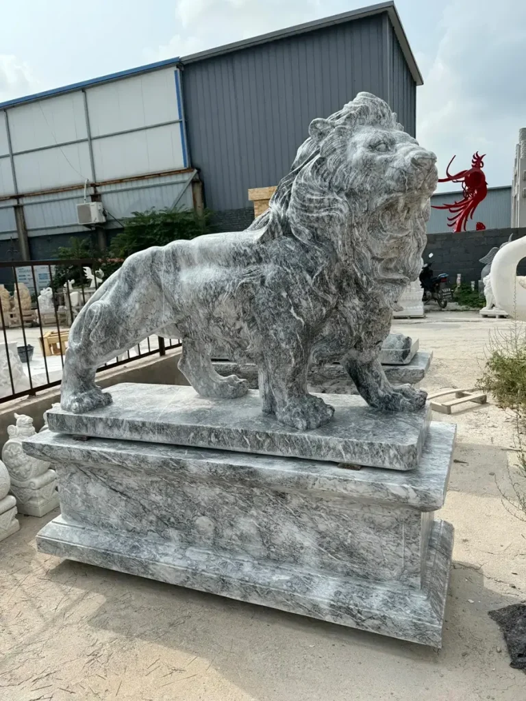 Side-front view of a majestic outdoor lion statue carved from heavily veined grey marble, standing securely on a matching pedestal. The premium natural stone allows for extremely fine detailing in the roaring facial features, perfect for achieving a classical estate look.