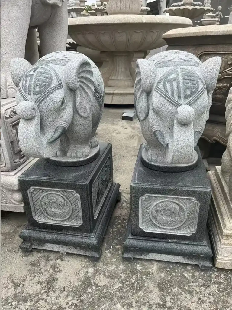Two grey stone elephant sculptures on black pedestals featuring the Chinese character for 'Blessing' (Fu) on their foreheads and their trunks pointing up. This upward gesture acts like a fountain releasing good fortune and positive energy into the environment.