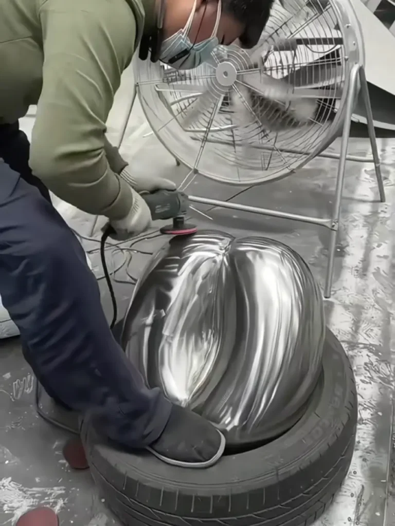 A worker using a power grinder to smooth the curved surfaces and weld seams of the assembled metal sculpture. This hands-on refinement is essential to the direct metal sculpture technique before final polishing.