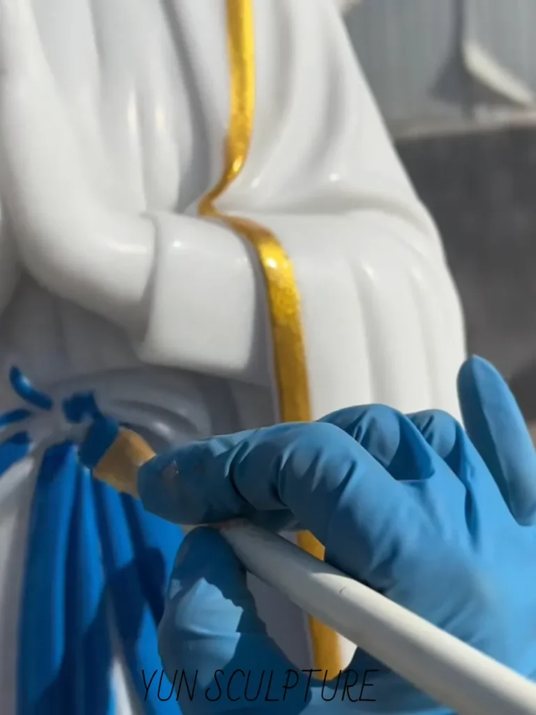 Extreme close-up of a gloved artisan meticulously hand-painting the distinct blue sash on a white marble Our Lady of Lourdes statue. Adhering rigidly to Saint Bernadette's eyewitness testimony, this precise brushwork creates a stark visual contrast against the brilliant white robe, fulfilling the strict iconographic constitution of the 1858 apparitions.