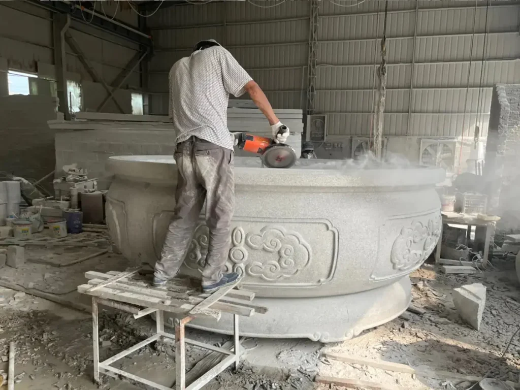 An artisan standing on a platform to polish the rim of a giant round granite planter, demonstrating the reinforced construction and structural stability of custom stone pieces.