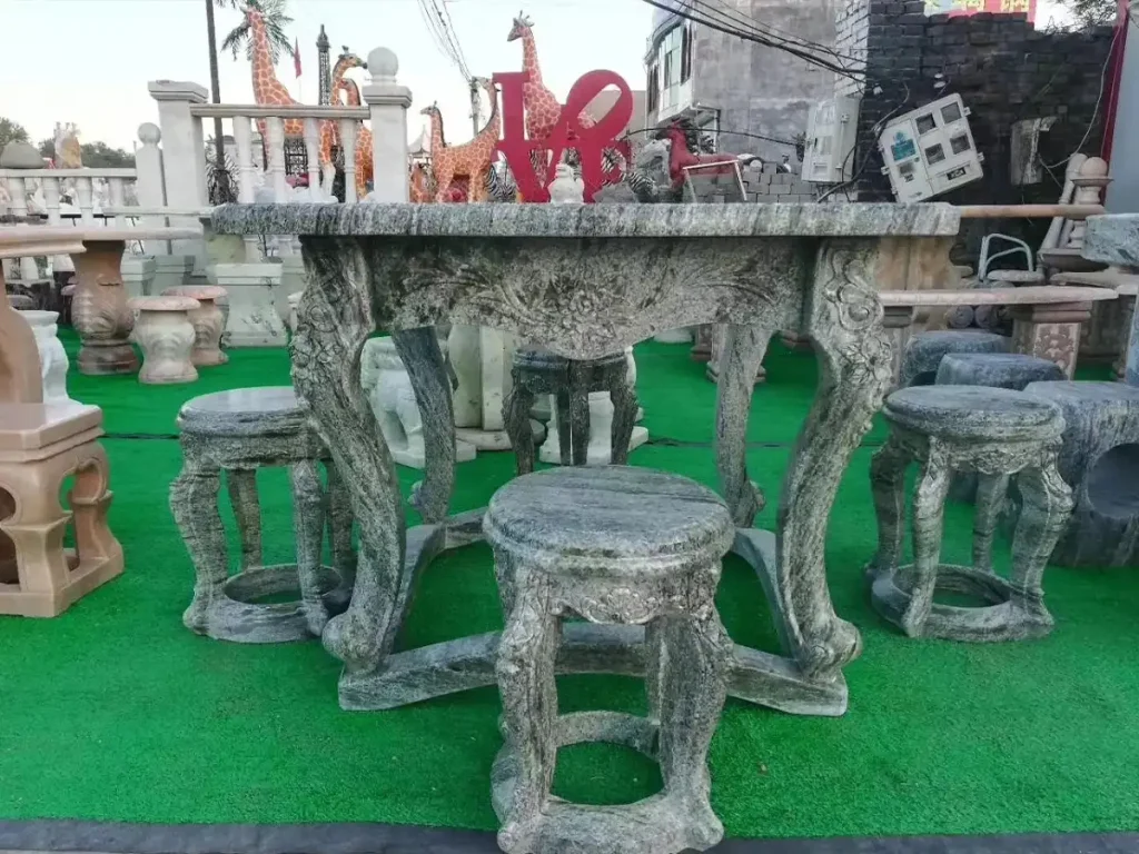 A heavy grey granite stone garden table set with intricately carved legs and matching drum stools. Displayed in a stone yard, its massive weight and natural texture establish a permanent visual and tactile boundary, ensuring this outdoor dining centerpiece will never shift with the wind.