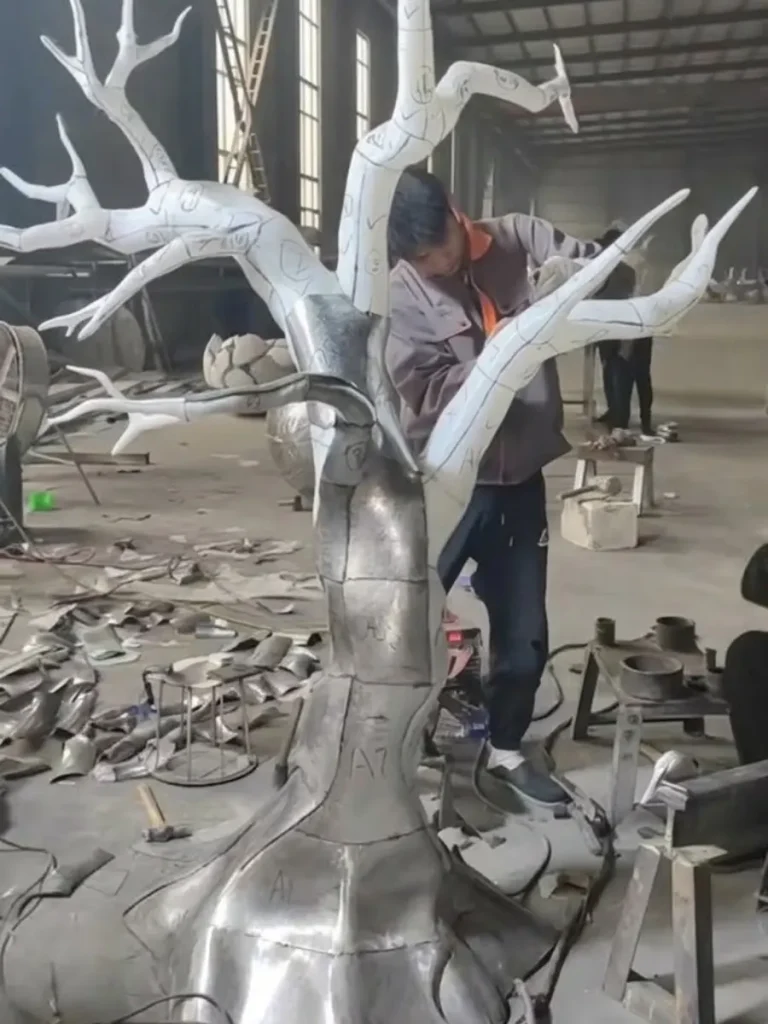 A craftsman meticulously fitting and welding curved stainless steel panels onto the tree structure. This hollow forging technique creates monumental visual mass and depth without the excessive weight of solid casting, balancing artistry with structural engineering.