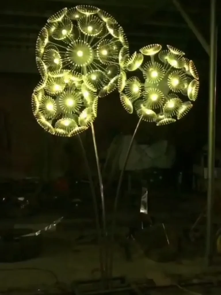 A cluster of three giant metal flower sculptures shaped like dandelions, glowing brightly with internal illumination in a darkened factory setting. These stunning illuminated artworks act as a "frozen garden," humanizing large spaces and creating an intimate, magical narrative when placed outdoors.