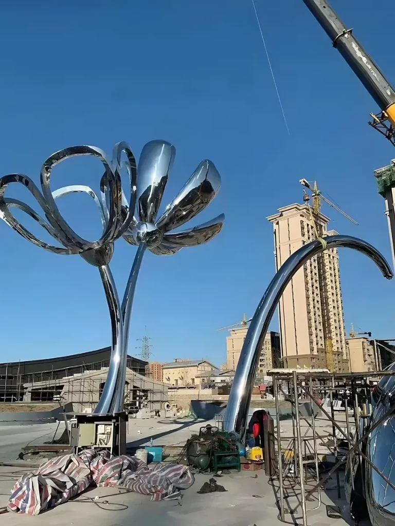 A heavy-duty crane positioning a giant, multi-ton stainless steel plant sculpture in an open plaza. This critical installation phase highlights that a pre-engineered concrete foundation and stainless steel expansion bolts are absolutely essential to resist uplift and lateral movement in extreme weather.