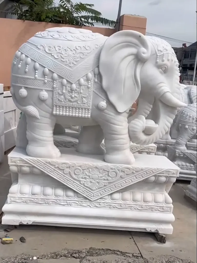 A detailed profile view of a life-size white marble elephant statue. The exquisitely carved ceremonial blanket and ornate headdress on the sculpture beautifully capture the revered royal status and rich historical symbolism of elephants across different civilizations.