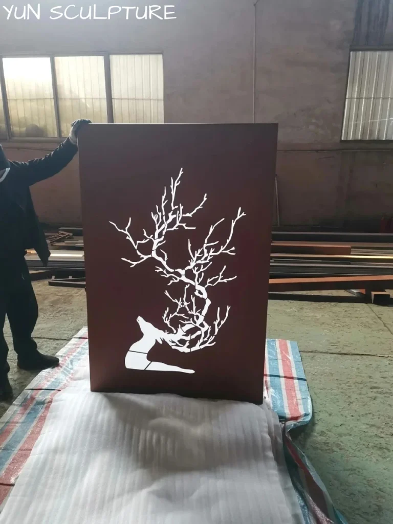 A worker holding a rusted iron wall sculpture with an intricate deer and branch cutout in a workshop, showcasing the openwork design that is essential for creating dynamic shadow play.