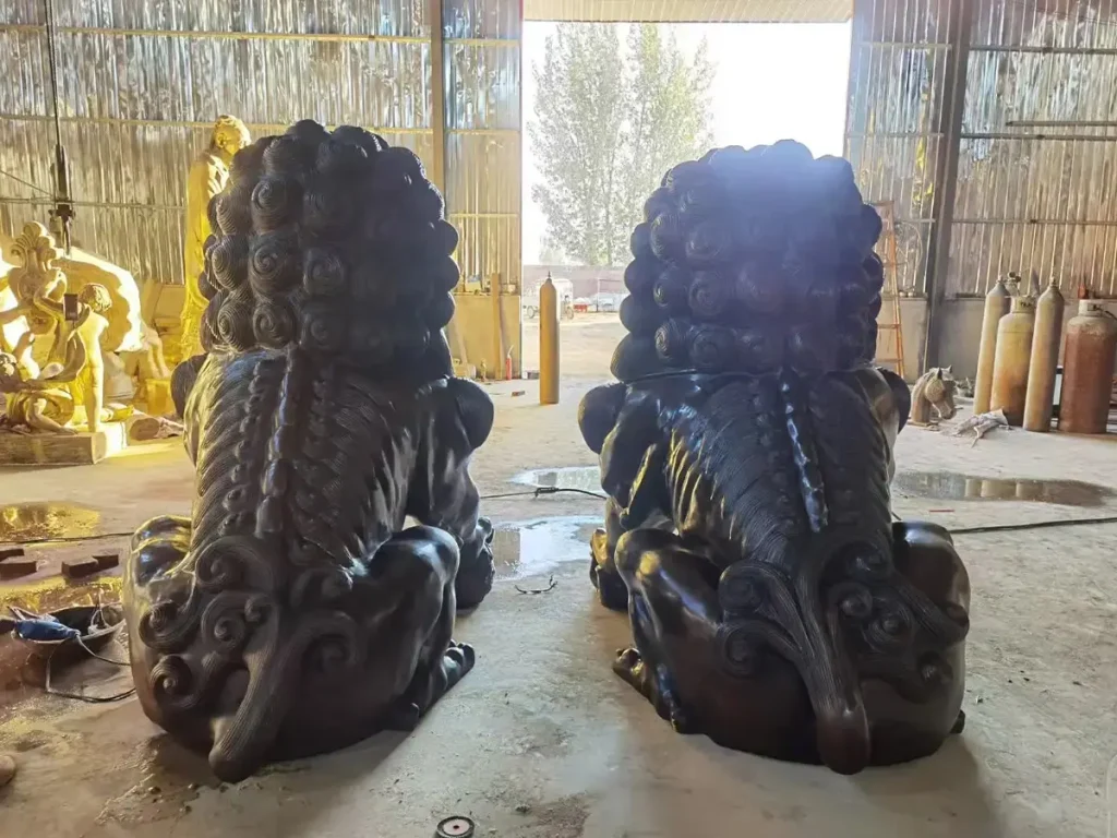 Rear view of two imposing, dark bronze estate lions inside a casting workshop, surrounded by foundry equipment. The detailed sculpting of their layered manes, spines, and curled tails highlights the imperial grandeur and exact proportions achieved by modern custom foundries.