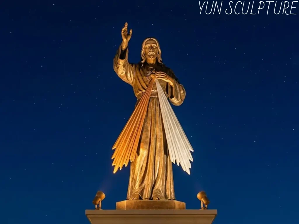 A large Divine Mercy bronze statue with signature red and pale rays, illuminated at night as a permanent outdoor monument installation.