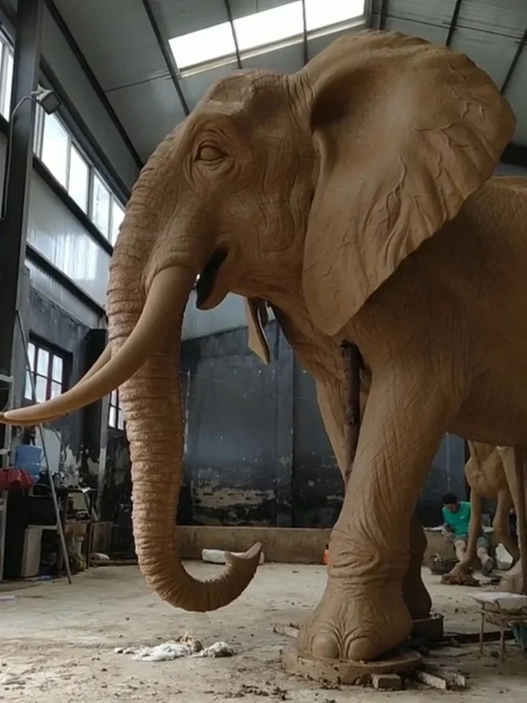 Diagonal view of a giant clay elephant model being meticulously sculpted. The sheer size of the artwork relative to the workshop environment highlights the emotional power of scale, proving how a carefully positioned life-sized elephant can transform an ordinary park into a space of majestic presence.