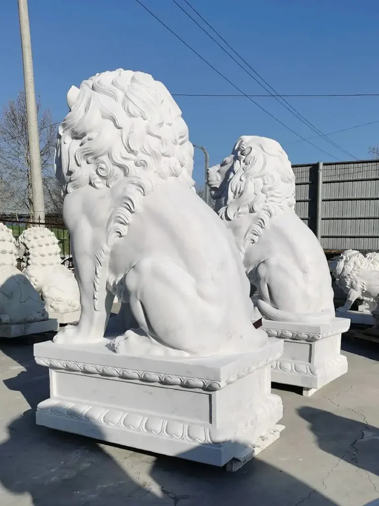 Side-angled view of two massive white marble lion statues waiting in a workshop yard. The deep physical profile and solid stone construction highlight the sheer volume required to ensure the sculptures do not get dwarfed by massive architectural structures or long driveways.