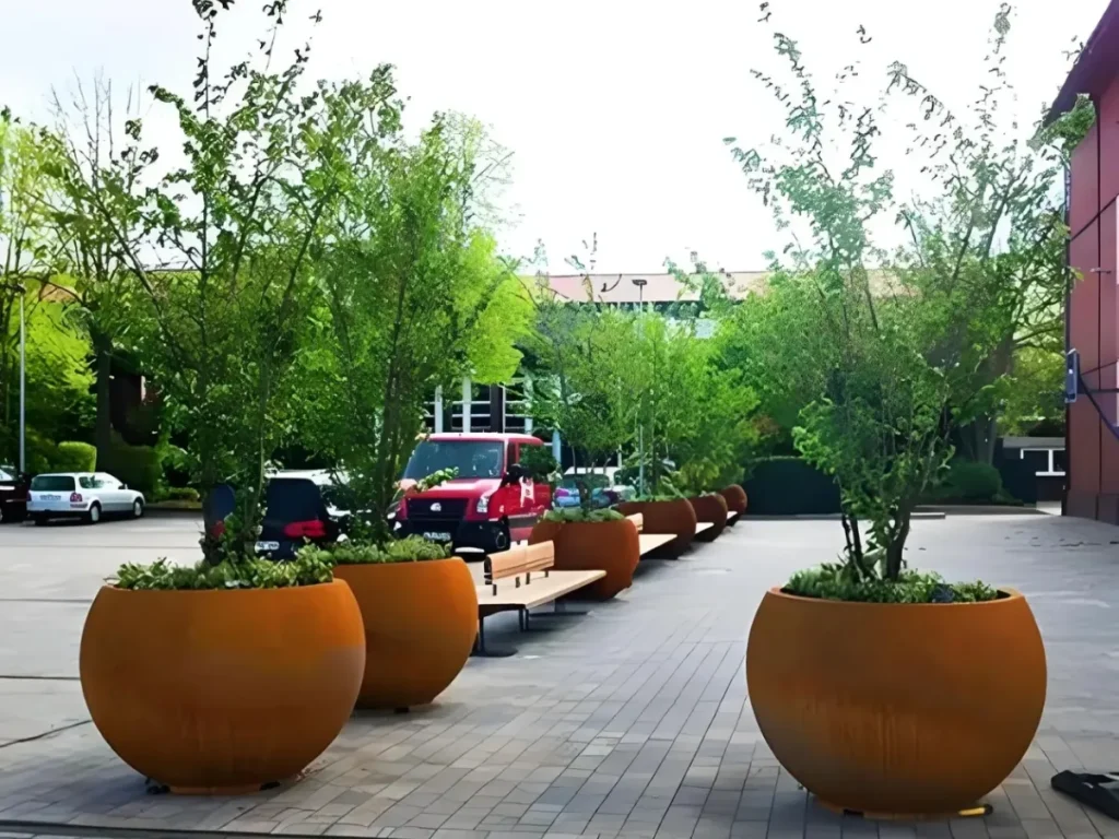 A row of massive, spherical corten steel planters holding vibrant green trees in a paved contemporary urban plaza. Showcasing a rich, earthy rust finish, these weathering steel planters perfectly illustrate the balance between industrial and organic design, anchoring the public space with undeniable architectural weight and a beautiful living patina.