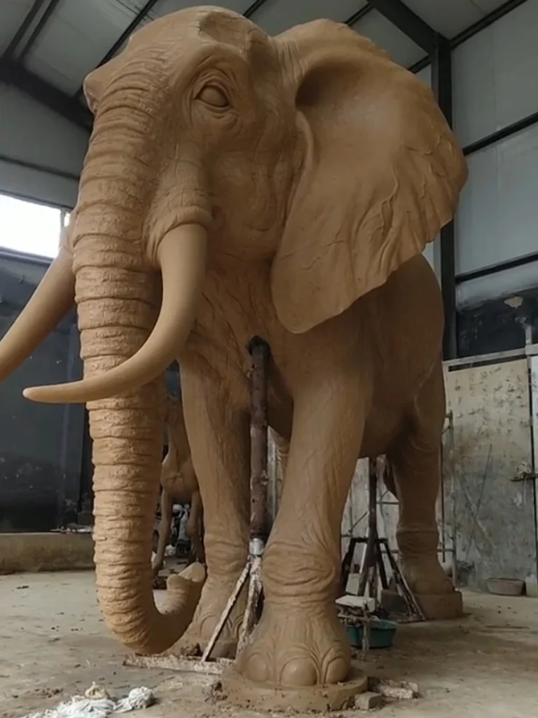 A massive life-size clay model of an elephant statue under construction in a sculptor's workshop. Supported by internal armatures and wooden props, this impressive prototype demonstrates the grand scale required for large outdoor elephant statues used as striking focal points in luxury estate gardens.