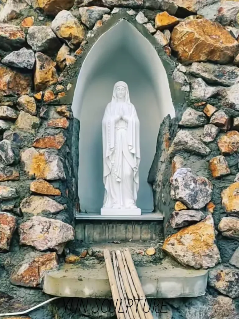 A full frontal view of a Marian grotto under construction, featuring a white marble statue inside a niche surrounded by heavily textured natural stone veneer over a reinforced core. The visible wooden ramp and unfinished masonry work perfectly illustrate the complex structural foundation and critical drainage slope engineering necessary to prevent ice expansion behind the sacred artwork.