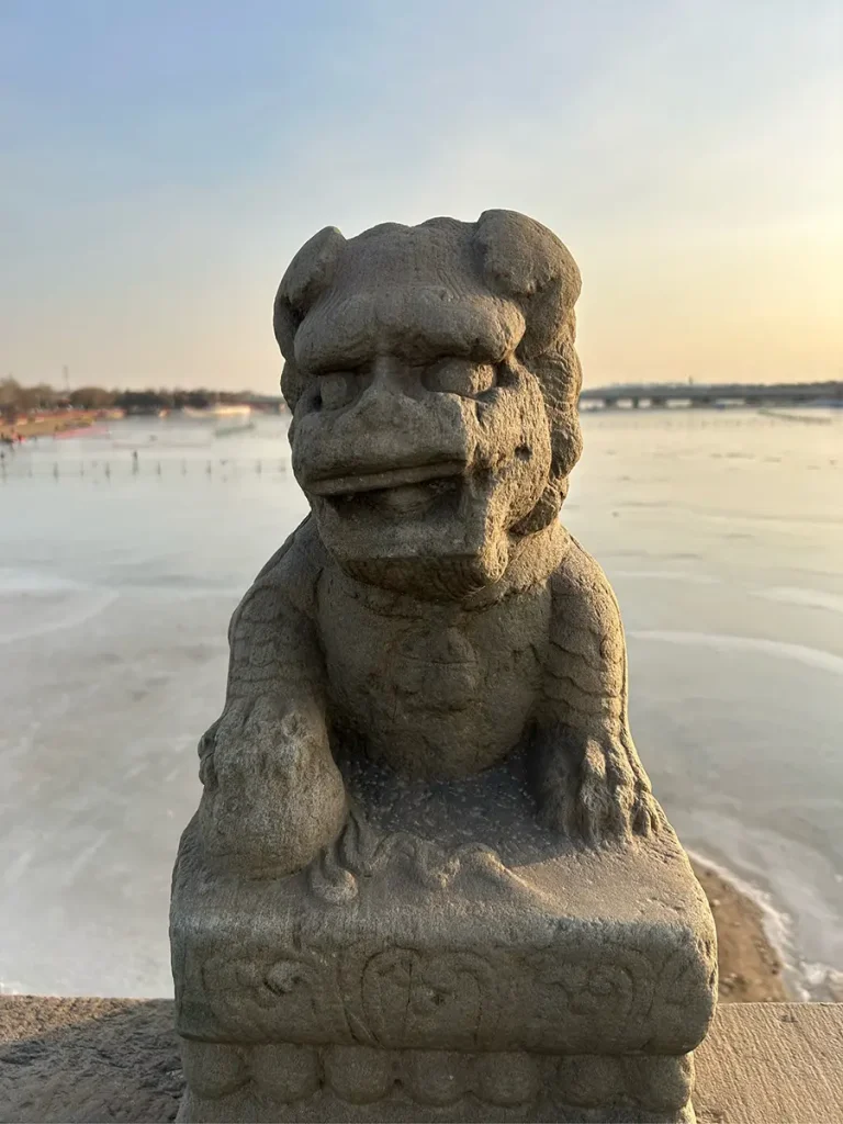 Front view of an ancient stone lion on the Marco Polo Bridge. The foo dog rests its right front paw firmly on the base, set against a beautiful backdrop of a partially frozen river under a soft sunset sky.