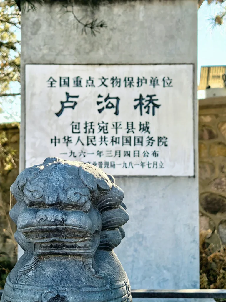 A small, detailed stone guardian lion sitting prominently in front of a stone stele. The stele marks the Lugou Bridge as a Major Historical and Cultural Site Protected at the National Level in China.