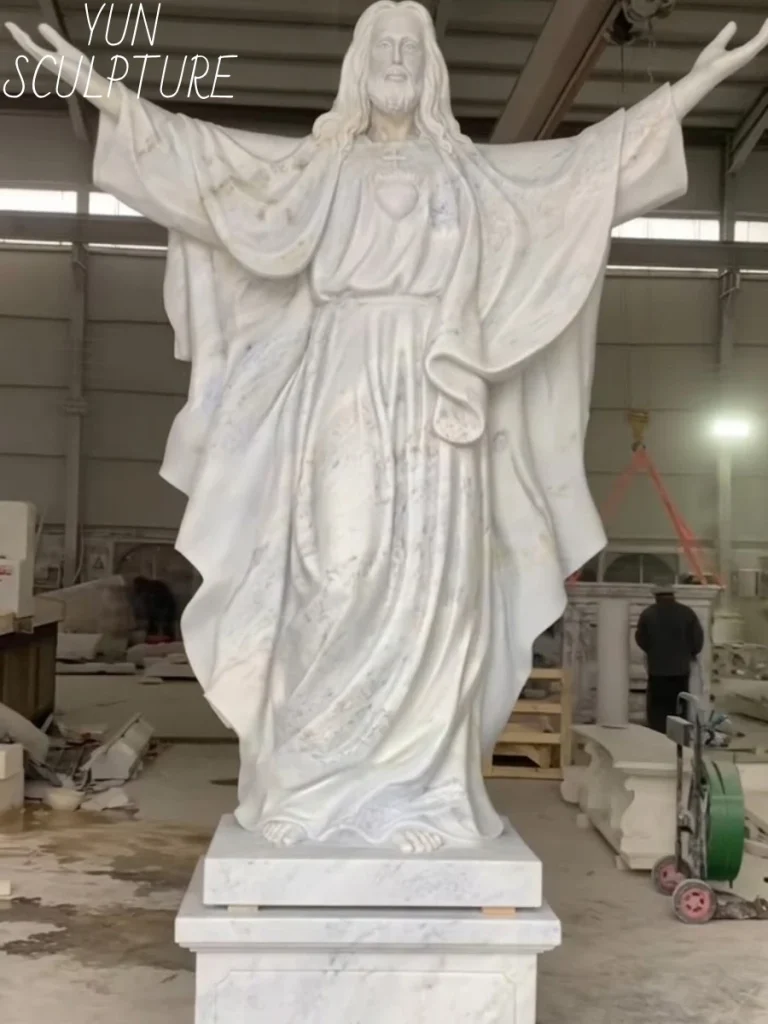A monumental, undeniably magnificent white marble jesus statue with outstretched arms standing in a workshop. Carved from premium white stone, this classical masterpiece is perfectly suited for climate-controlled indoor sanctuaries where its brilliant surface is protected from harsh outdoor elements.