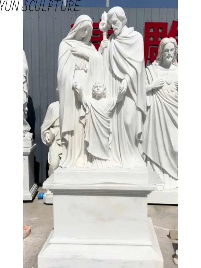 A white marble Jesus Mary and Joseph statue on a stone pedestal, designed as a welcoming 'open circuit' composition for a church plaza.