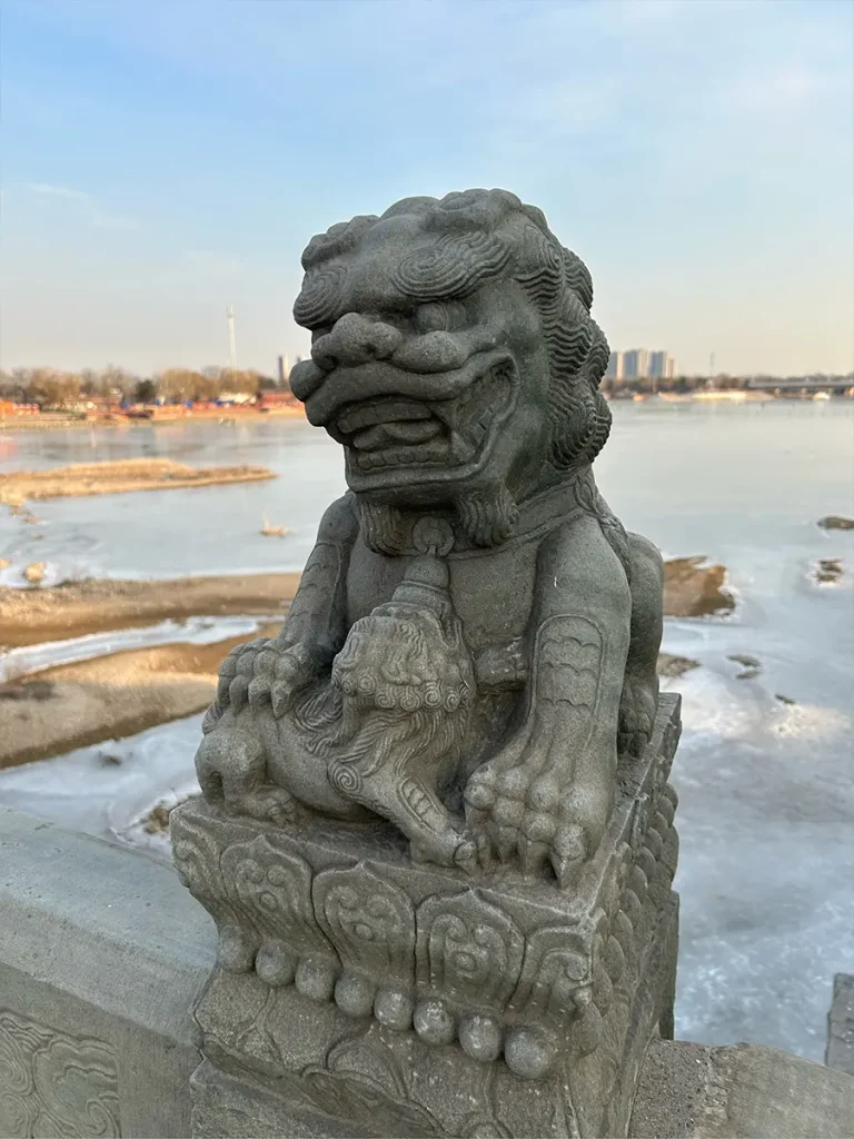 A female stone foo dog on the Lugou Bridge with her front paw gently resting on a small, finely carved cub. The statue sits on a decorative bridge pillar with a frozen, icy river and distant skyline in the background.