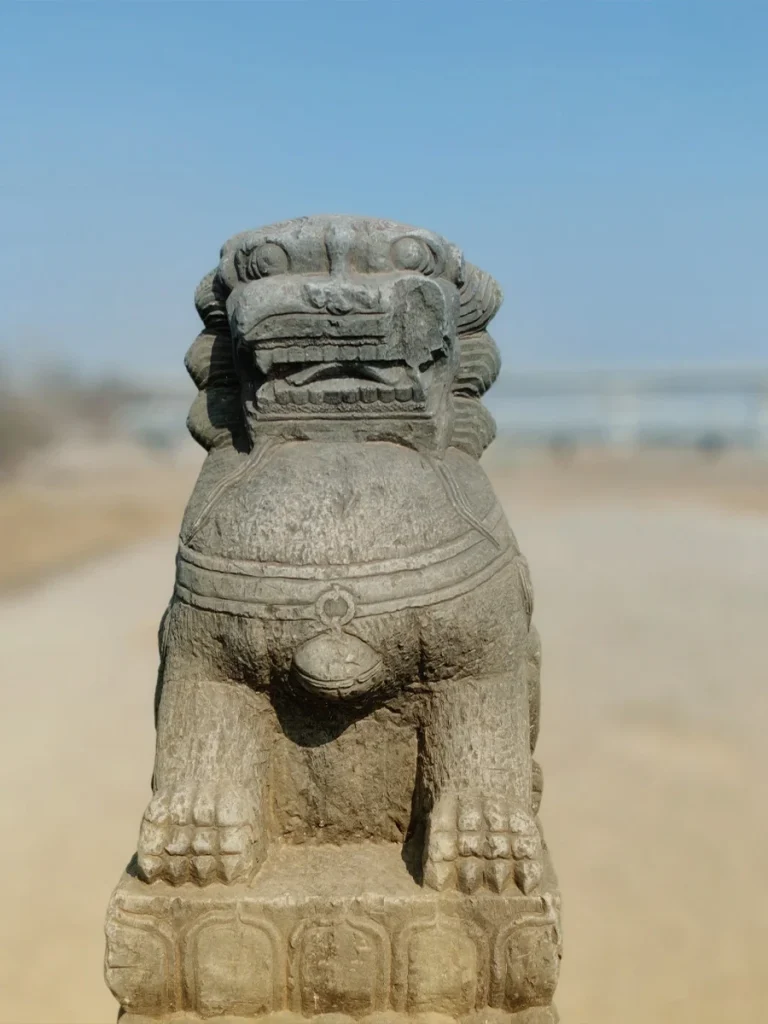 Close-up front view of a heavily weathered ancient stone foo dog on the Lugou Bridge. The lion's facial features and mane have been smoothed by centuries of wind and rain, presented against a blurred background of the riverbed.
