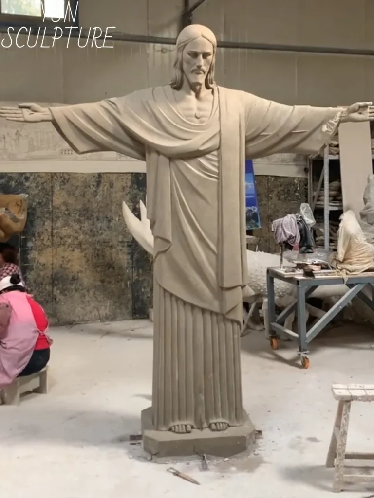 A detailed view of a masterfully hand-sculpted clay prototype for a bronze Jesus statue, highlighting the profound weight of the draped robe and the tension in the outstretched hands. Trusting the human eye over digital models ensures these crucial anatomical details communicate triumph and resurrection.