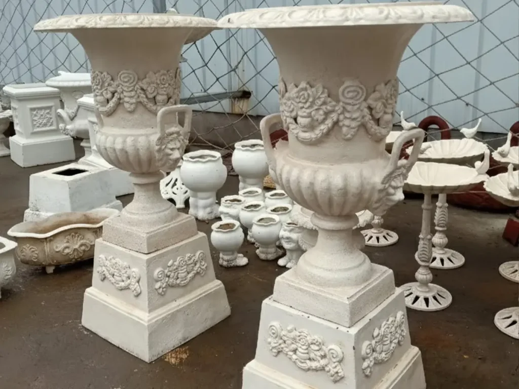 A matching pair of highly ornate white cast iron urns and pedestals displayed in a workshop. Elevating the planters on these architectural bases adds height, authority, and formality, transforming a simple garden pot into a valuable statement piece.