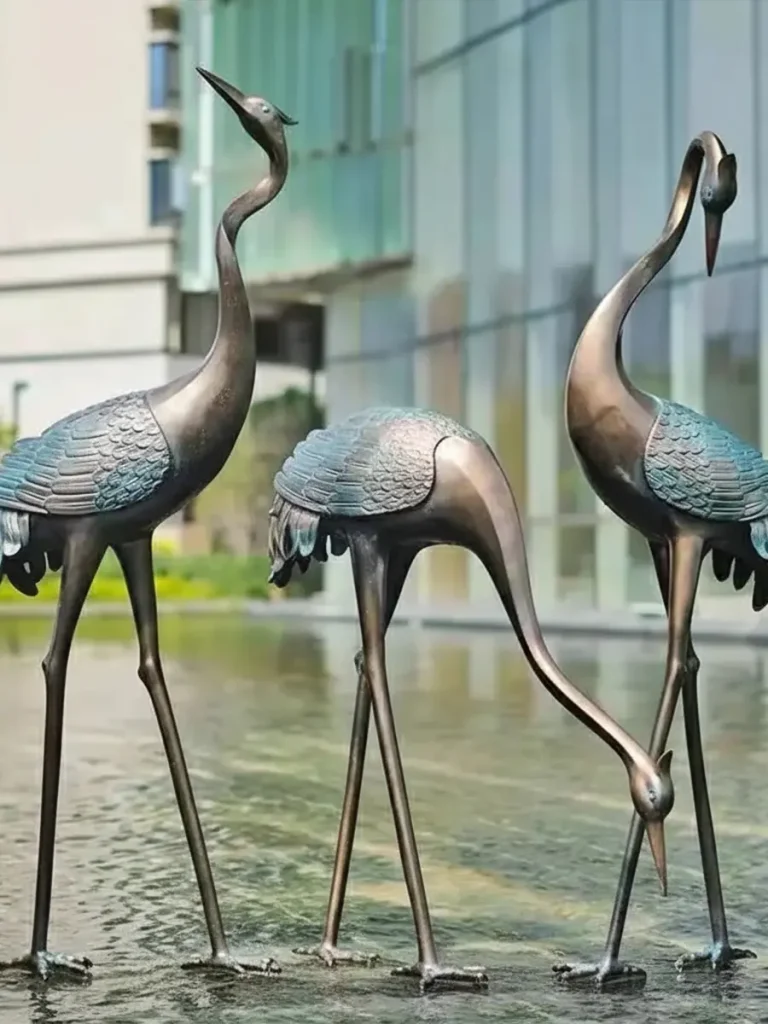 An elegant group of bronze sculptures standing gracefully in a shallow water feature outside a modern building. This beautiful metal crane sculpture set highlights the iconic patina copper finish on its wings, bringing an organic warmth and a sense of cultural continuity to the landscape design.