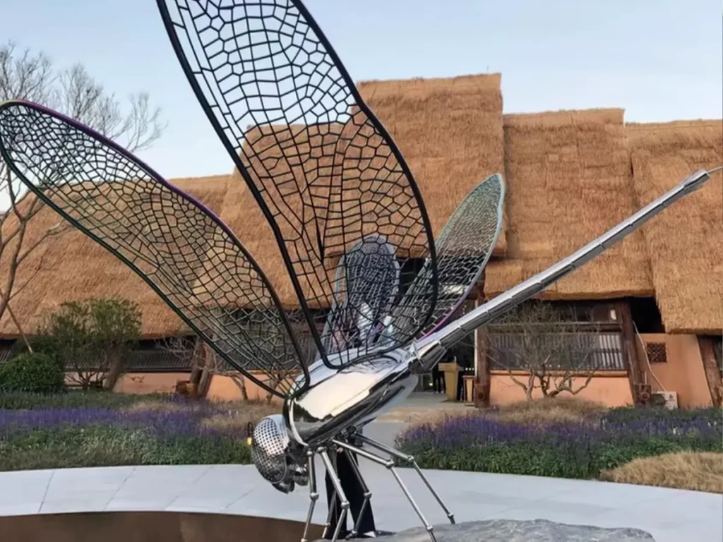 A giant, mirror-polished metal dragonfly sculpture installed in a modern outdoor landscape. Featuring beautifully crafted wire-mesh wings, this stunning modern metal sculpture acts as a poetic accent, animating the space and introducing gentle movement to the quiet surroundings.