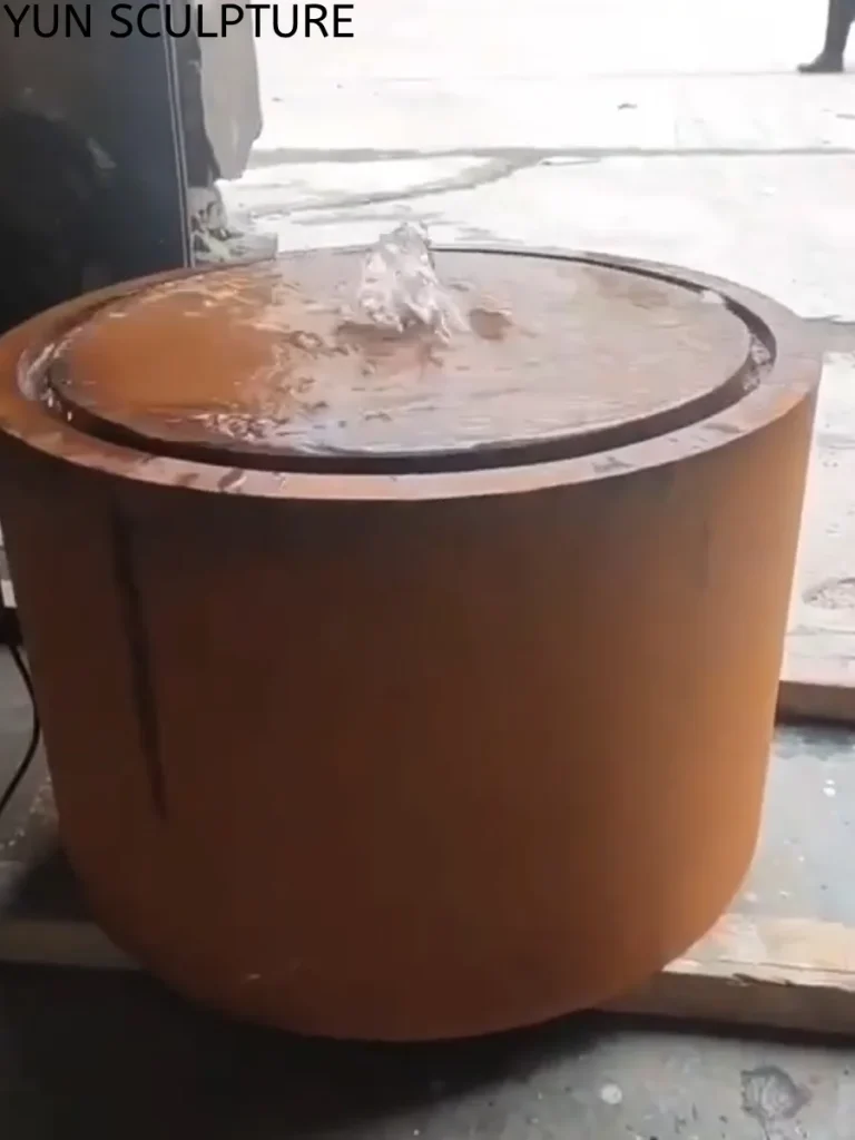 Close-up of a modern round metal outdoor fountain crafted from weathering steel during a factory test. The water cascades perfectly into a recessed rim, keeping the outer walls free from stagnant water. This specific design respects the wet-dry cycles necessary to stabilize the Corten steel alloy and prevent structural failure.