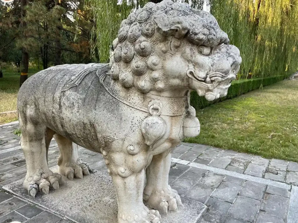 A majestic standing stone Foo Dog statue at the Ming Dynasty Tombs. This massive historical sculpture is depicted standing on all four legs, showcasing natural weathering on its smooth stone back, distinct tight curls on its head, and large decorative carved bells hanging across its chest.