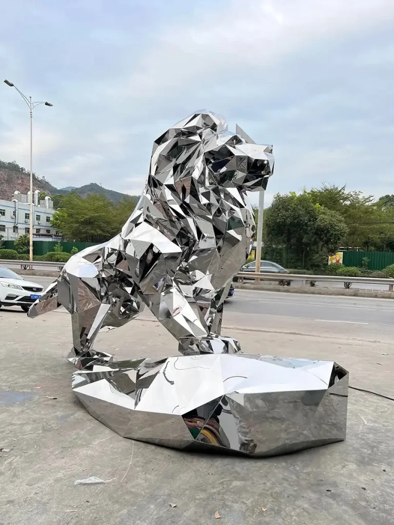 A large, modern mirror-polished 316 stainless steel lion statue displayed outdoors. The sculpture acts as a dynamic visual element, with its highly reflective geometric facets mirroring the sky, trees, and street, perfectly integrating the guardian symbol with modern design language.