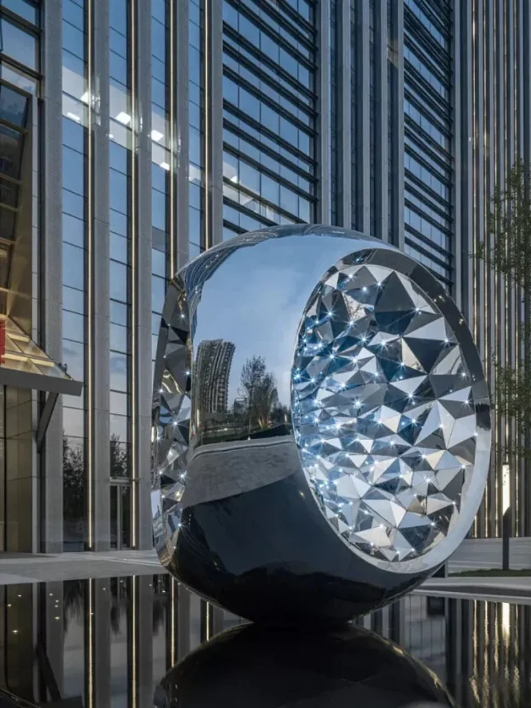A striking modern metal sculpture featuring a smooth mirror-polished exterior and a highly complex, faceted geometric interior. Placed on a reflective water surface in a corporate campus, this abstract artwork acts as a powerful visual anchor, perfectly illustrating the profound dialogue between the surrounding architectural structure and artistic intervention.