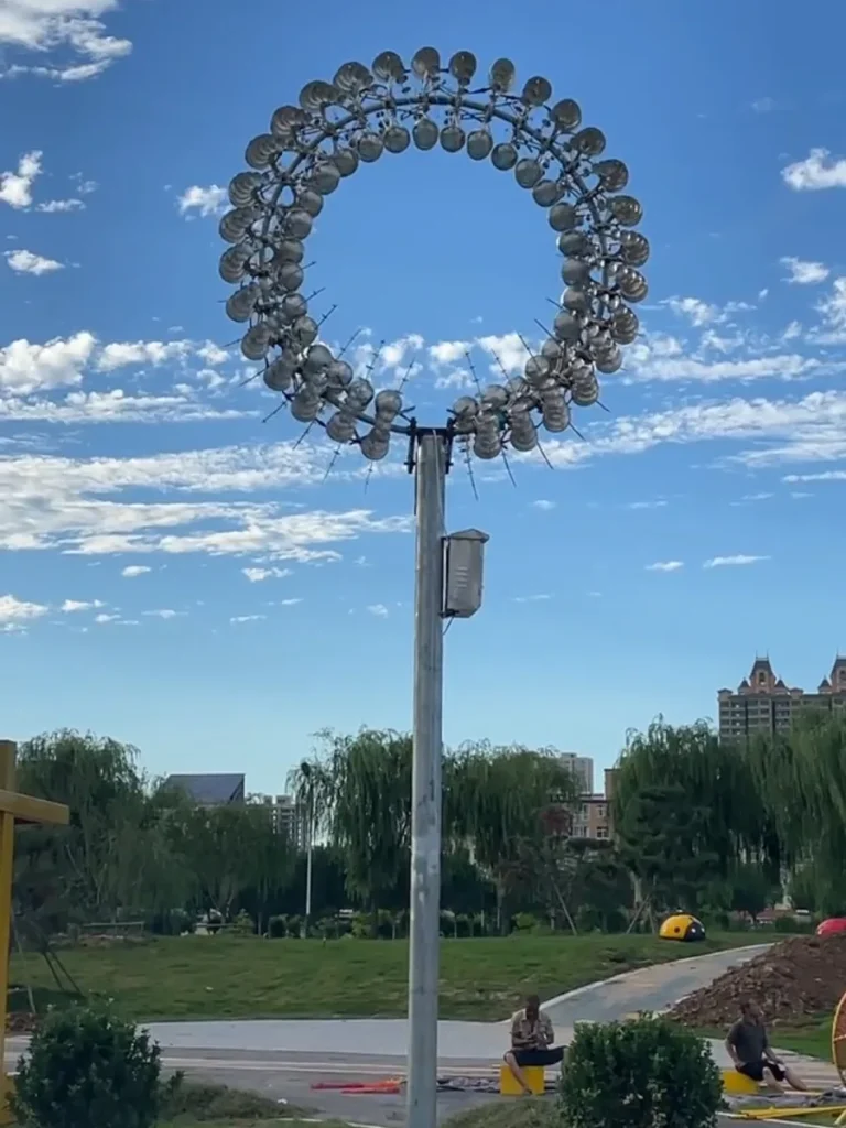 A modern wind sculpture kinetic artwork beginning to spin gently in an outdoor landscape. Acting as an instrument of nature, the reflective metal elements catch the passing breeze, beautifully interrupting the stillness of the lawn and providing a captivating visualization of wind.