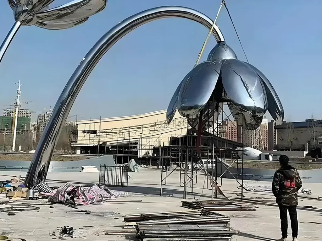 A monumental mirror-polished stainless steel plant sculpture being installed in an outdoor plaza. Capturing a single perfect moment of growth, this towering floral form features a flawless 8K mirror polish that creates the "Liquid Silver" effect. By brilliantly reflecting the blue sky and surrounding architecture, the outdoor stainless steel sculpture demonstrates how fleeting nature is masterfully transformed into a permanent, eternal focal point.