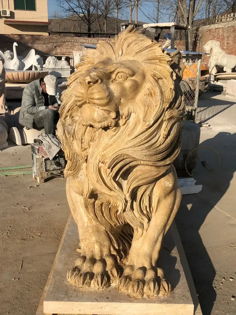 Direct front view of a highly detailed, antique-style yellow stone lion sculpture. The intense, majestic gaze directly engages approaching visitors, while the visible chest muscles and deep shadows around the eyes highlight the sculptor's mastery of real lion anatomy.